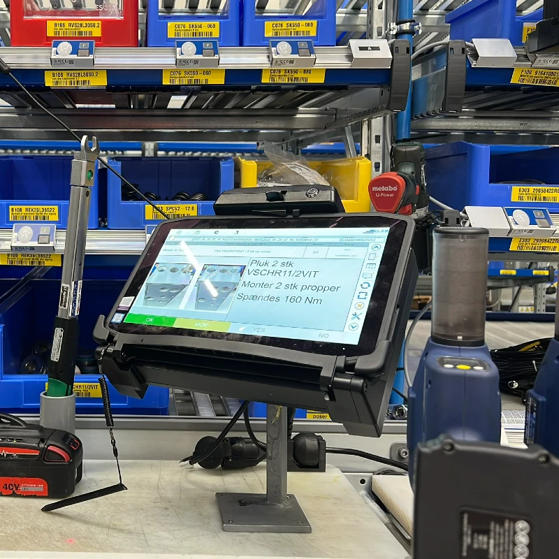 Tablet on a stand displaying instructions in a warehouse with blue bins and tools in the background.