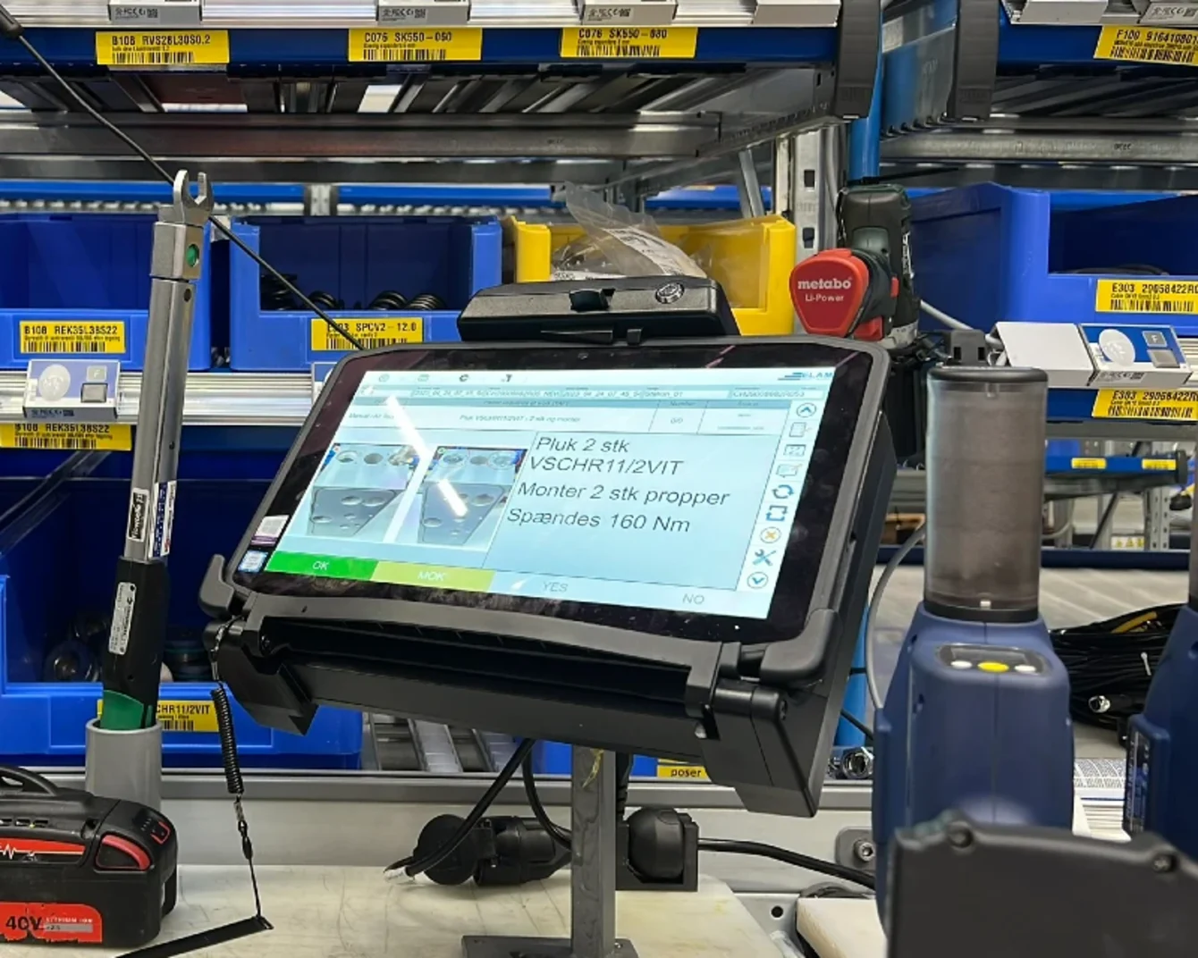 Tablet on a stand displaying instructions in a warehouse with blue bins and tools in the background.
