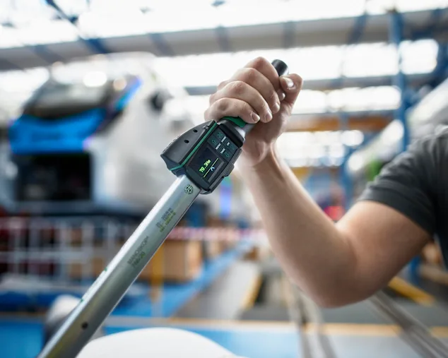 Person using digital torque wrench. Torque is visible on small display. The blurred background shows a train in a railway construction factory.