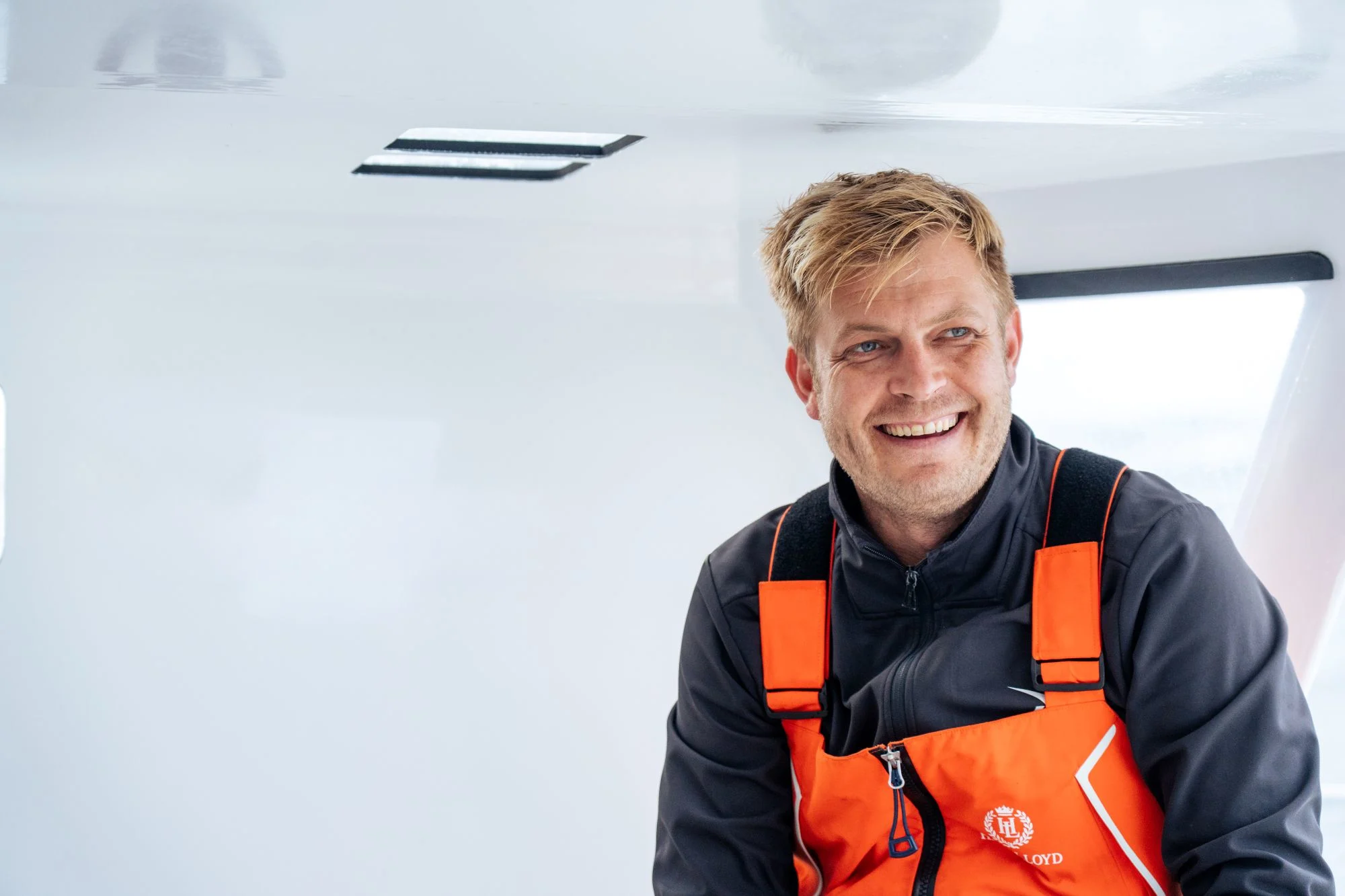 Smiling Oliver Heer in an orange and black life vest aboard a white boat with windows visible in background.