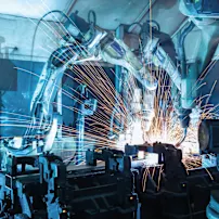Industrial robotic arms welding metal with bright orange sparks flying in a blue-lit factory setting.