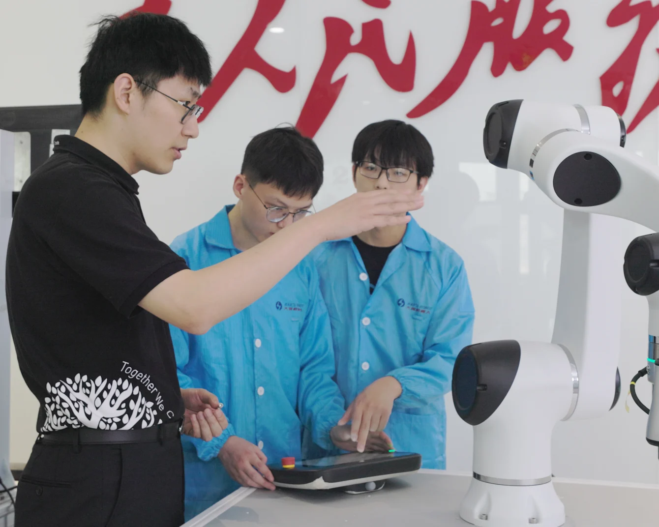 Three people in black and blue shirts examining a white robotic arm on a table in a lab setting.