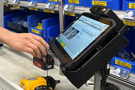 A hand interacts with a tablet on a stand in a workshop, surrounded by tools and blue storage bins with labels.