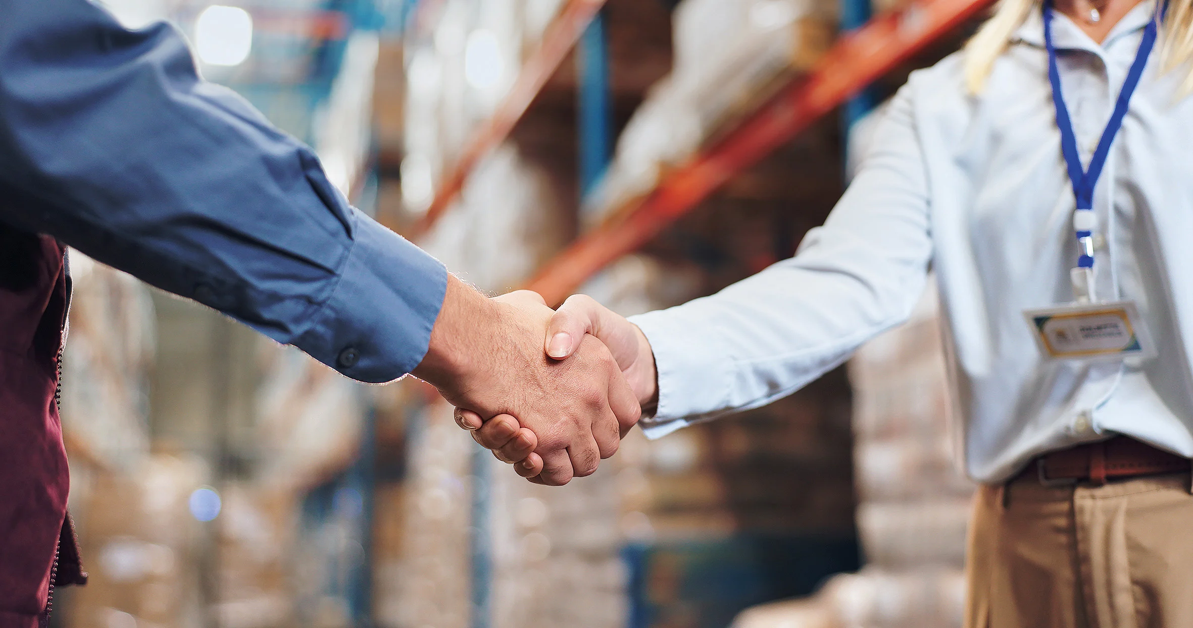 两个人在仓库握手,一个穿蓝色衬衫,另一个穿白色衬衫并佩戴名牌。