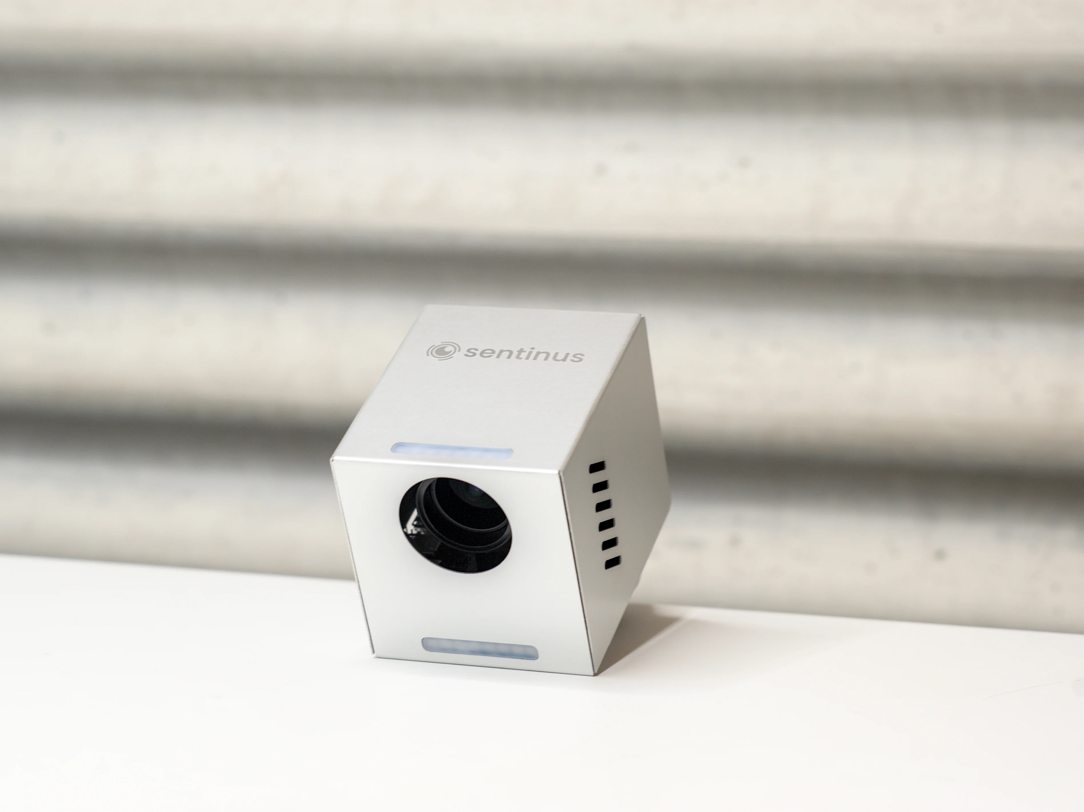 Silver cube-shaped device with a lens, labeled "sentinus," on a white surface against a textured gray background.