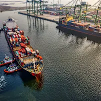 Cargo ship with colorful containers docked at a port, assisted by tugboats, with cranes and stacked containers in the background.