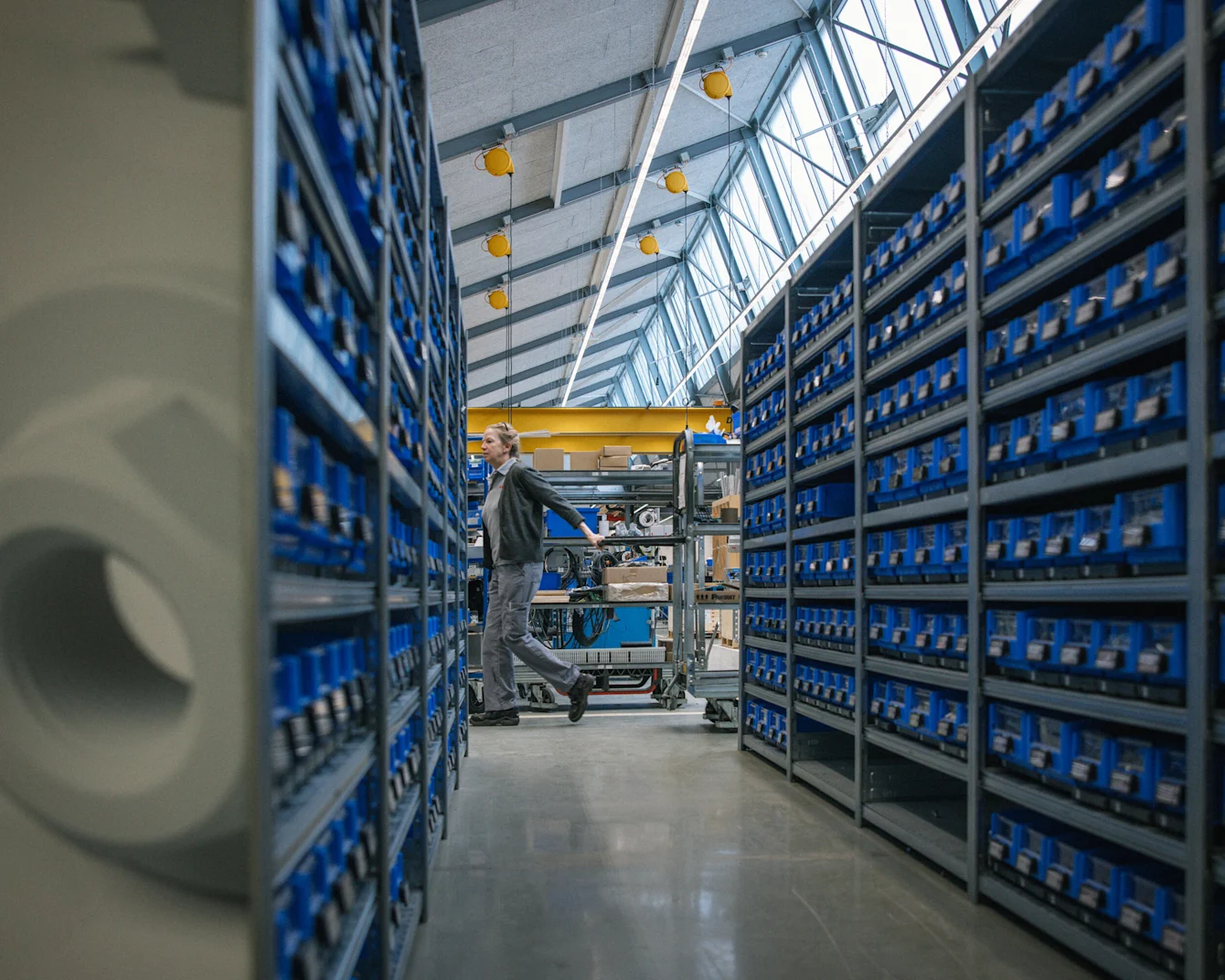 Worker pushing cart through modern warehouse aisle with tall blue storage shelves under glass ceiling with yellow lights.