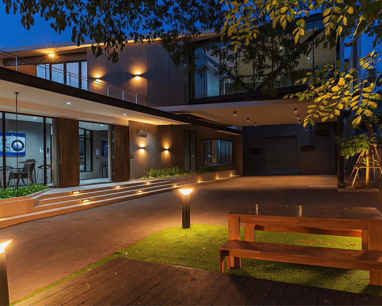 Modern house exterior at dusk with warm lighting, large windows, and a wooden bench on a grassy area, surrounded by trees.