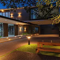 Modern house exterior at dusk with warm lighting, large windows, and a wooden bench on a grassy area, surrounded by trees.
