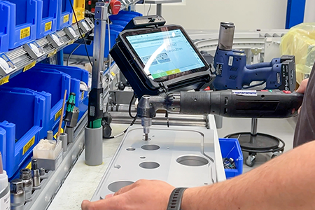 Person using a power tool on a metal panel, with a tablet displaying instructions nearby, surrounded by blue bins.