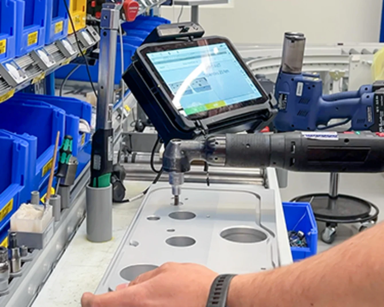 Person using a power tool on a metal panel, with a tablet displaying instructions nearby, surrounded by blue bins.