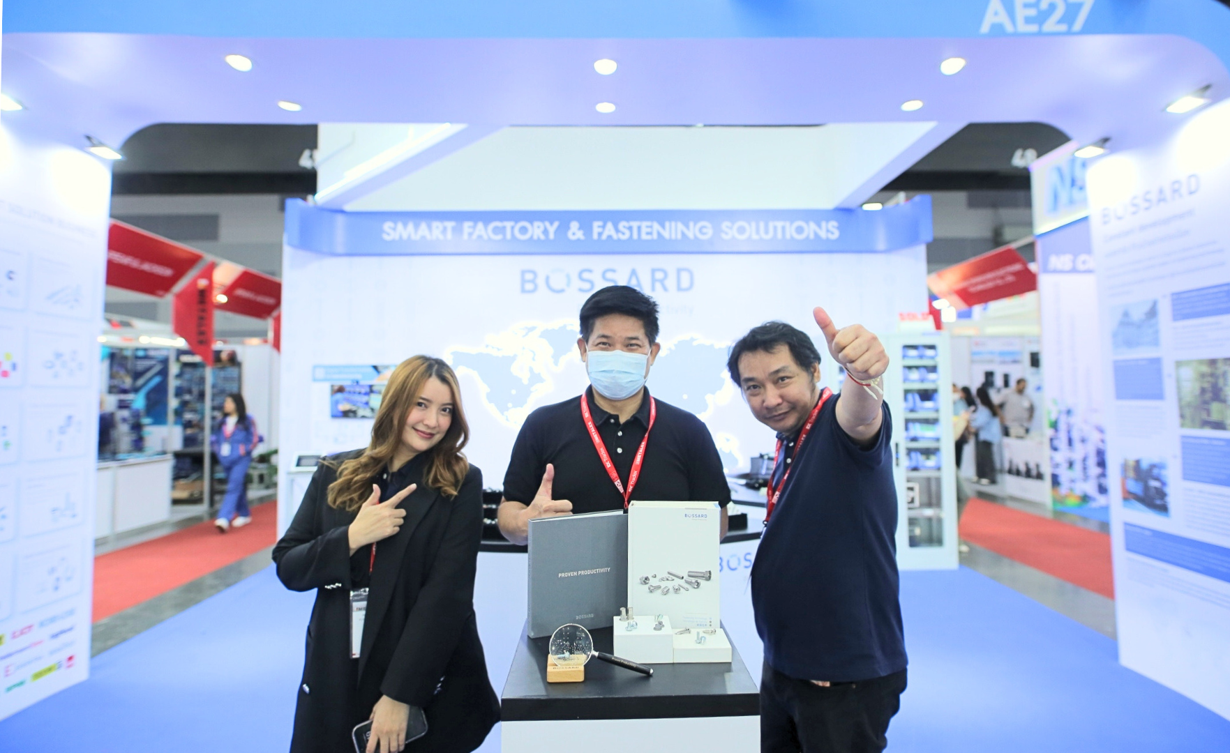Three Bossard employees at a trade fair stand in front of a booth marked "Smart Factory & Fastening Solutions," featuring Bossard's branding.