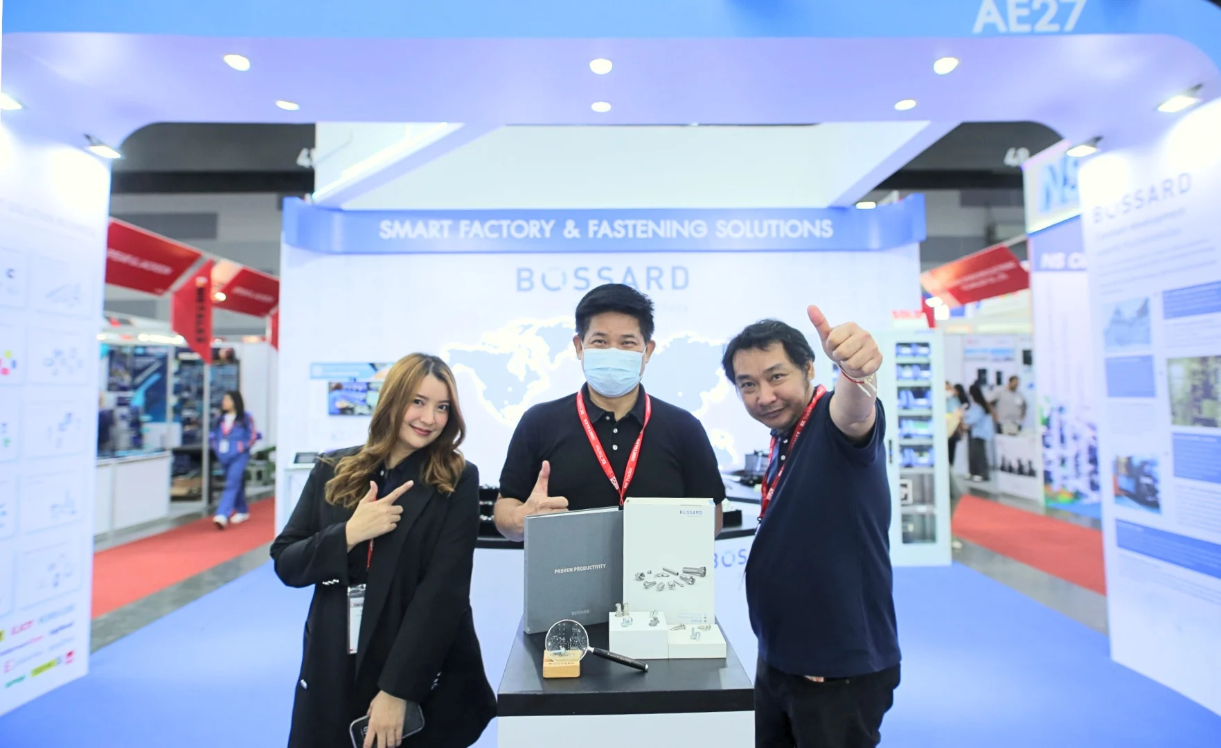 Three Bossard employees at a trade fair stand in front of a booth marked "Smart Factory & Fastening Solutions," featuring Bossard's branding.