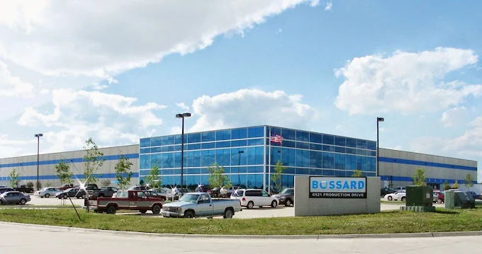 Large industrial building with blue-tinted windows, surrounded by parked cars and a sign reading "Bossard." Cloudy sky above.