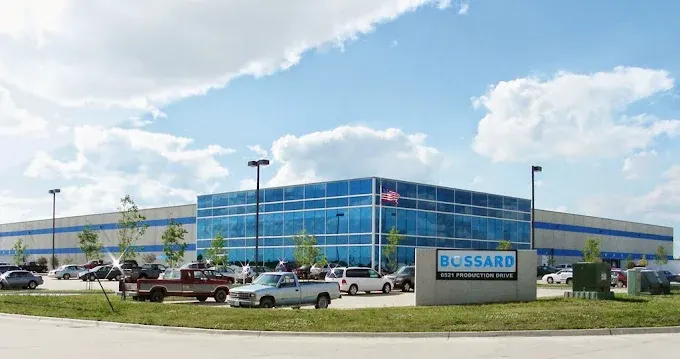 Large industrial building with blue-tinted windows, surrounded by parked cars and a sign reading "Bossard." Cloudy sky above.