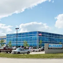 Large industrial building with blue-tinted windows, surrounded by parked cars and a sign reading "Bossard." Cloudy sky above.