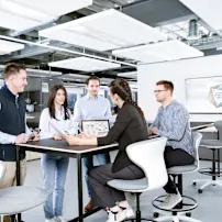 People around a table discussing a technical drawing in an industrial looking office. Key visual for Assembly Technology Expert Service only.