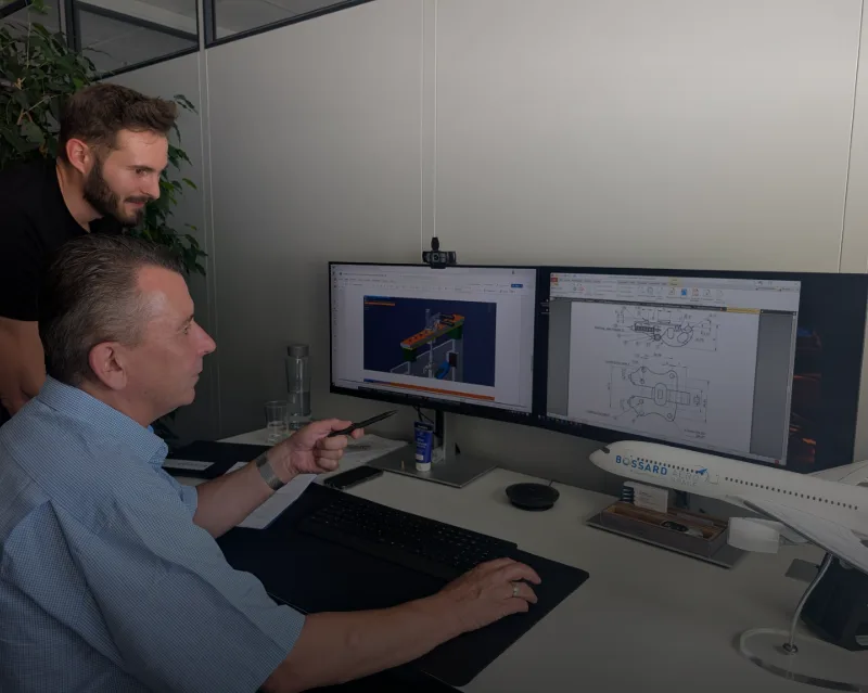 Two men reviewing CAD engineering drawings and 3D models on dual computer monitors in a modern office workspace.