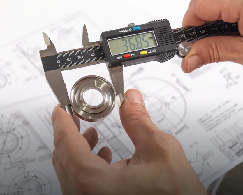 A pair of hands gripping a caliper to measure a metal bearing above technical drawings and specifications.