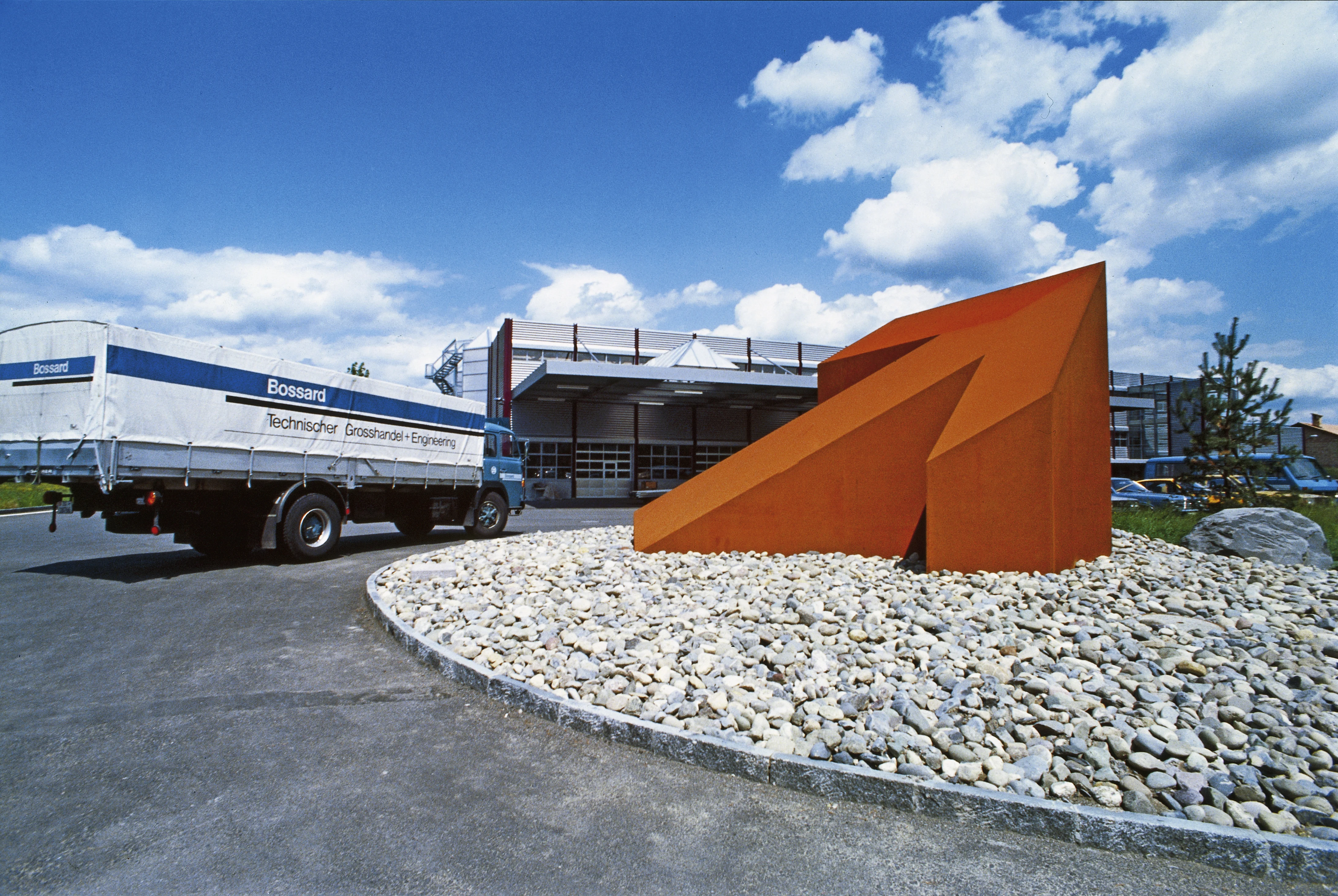 历史图片：一个大型橙色箭头雕塑位于环岛，背景中有Bossard柏中的仓库一辆卡车。