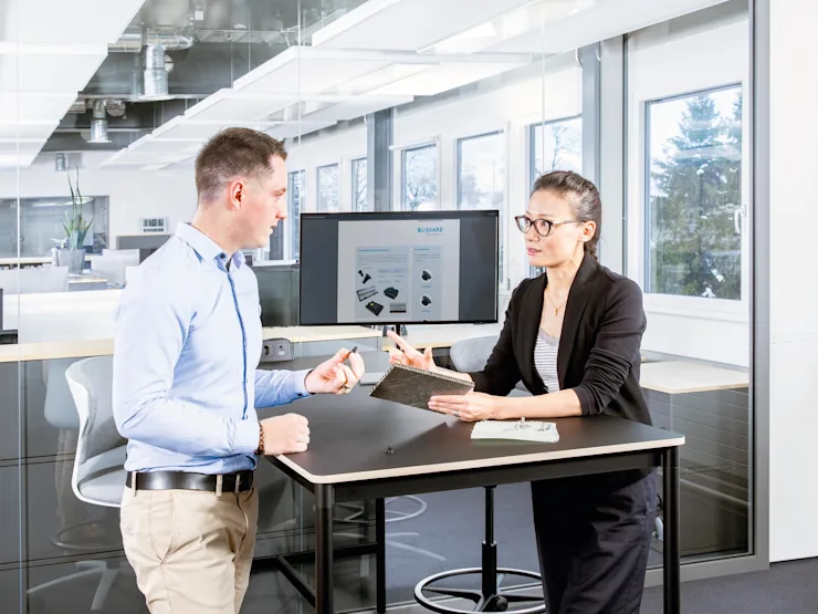Two people at a desk in a modern office discuss fastening a composite material; one holds a sandwich panel, the other a fastener.