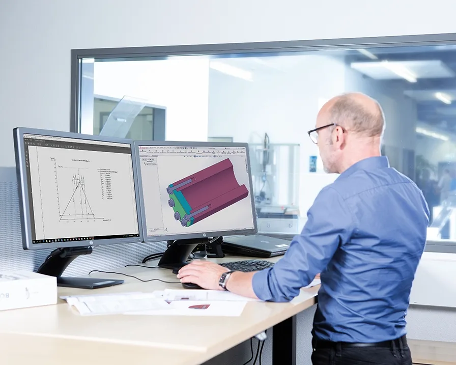 A man in a blue shirt works on a technical drawing at a standing desk on a computer