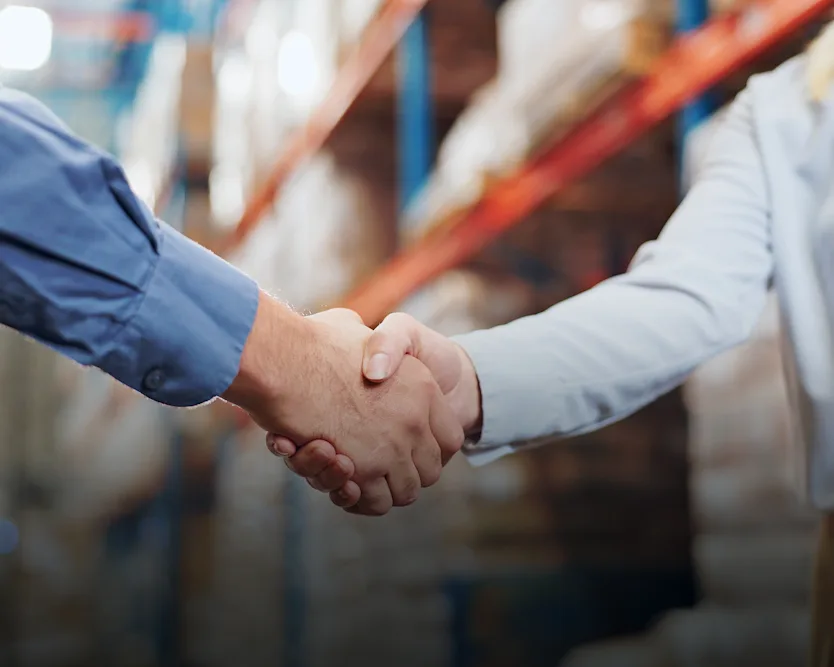 Two people shaking hands in a warehouse, one wearing a blue shirt, the other with a name badge, shelves in the background.