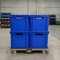 Blue plastic storage bins stacked on a smart pallet system in a warehouse with shelving and yellow safety posts on the floor.