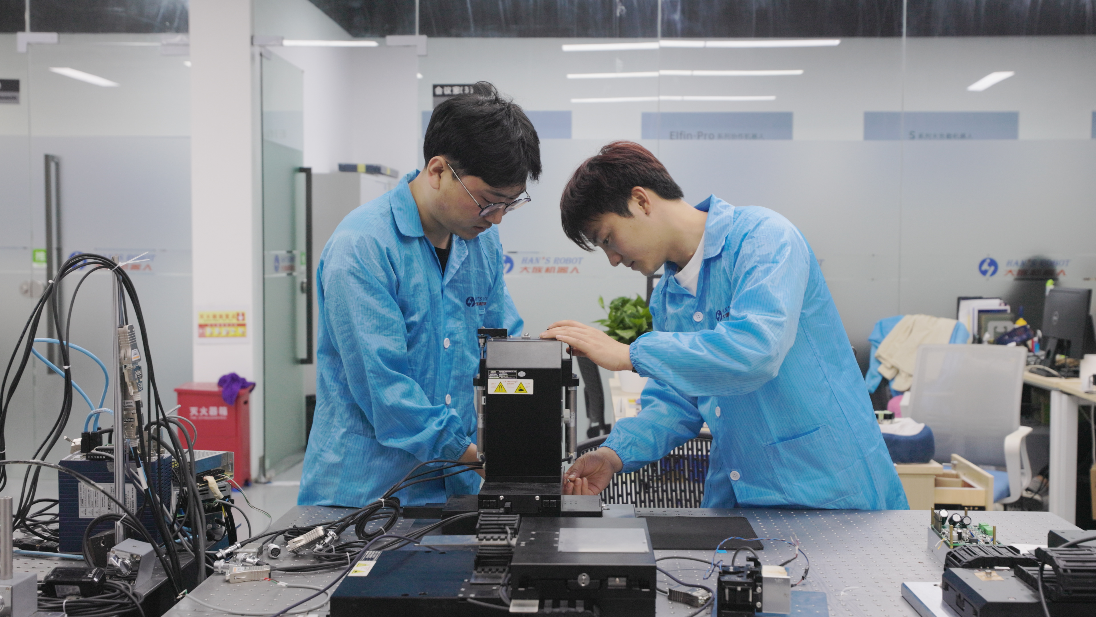 Two technicians in blue lab coats examining electronic equipment on a workbench in a modern laboratory setting.
