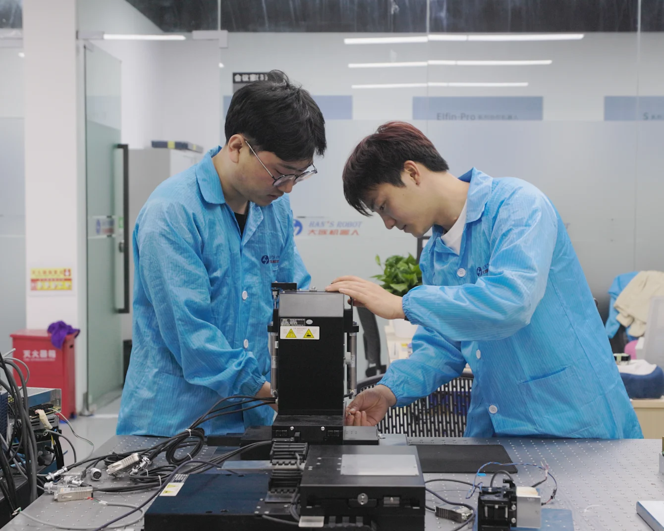 Two technicians in blue lab coats examining electronic equipment on a workbench in a modern laboratory setting.