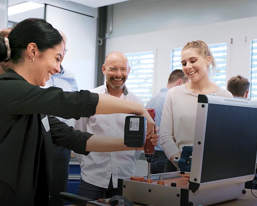 A group of people stands around a workbench, using tools and a cordless screwdriver on the workbench.