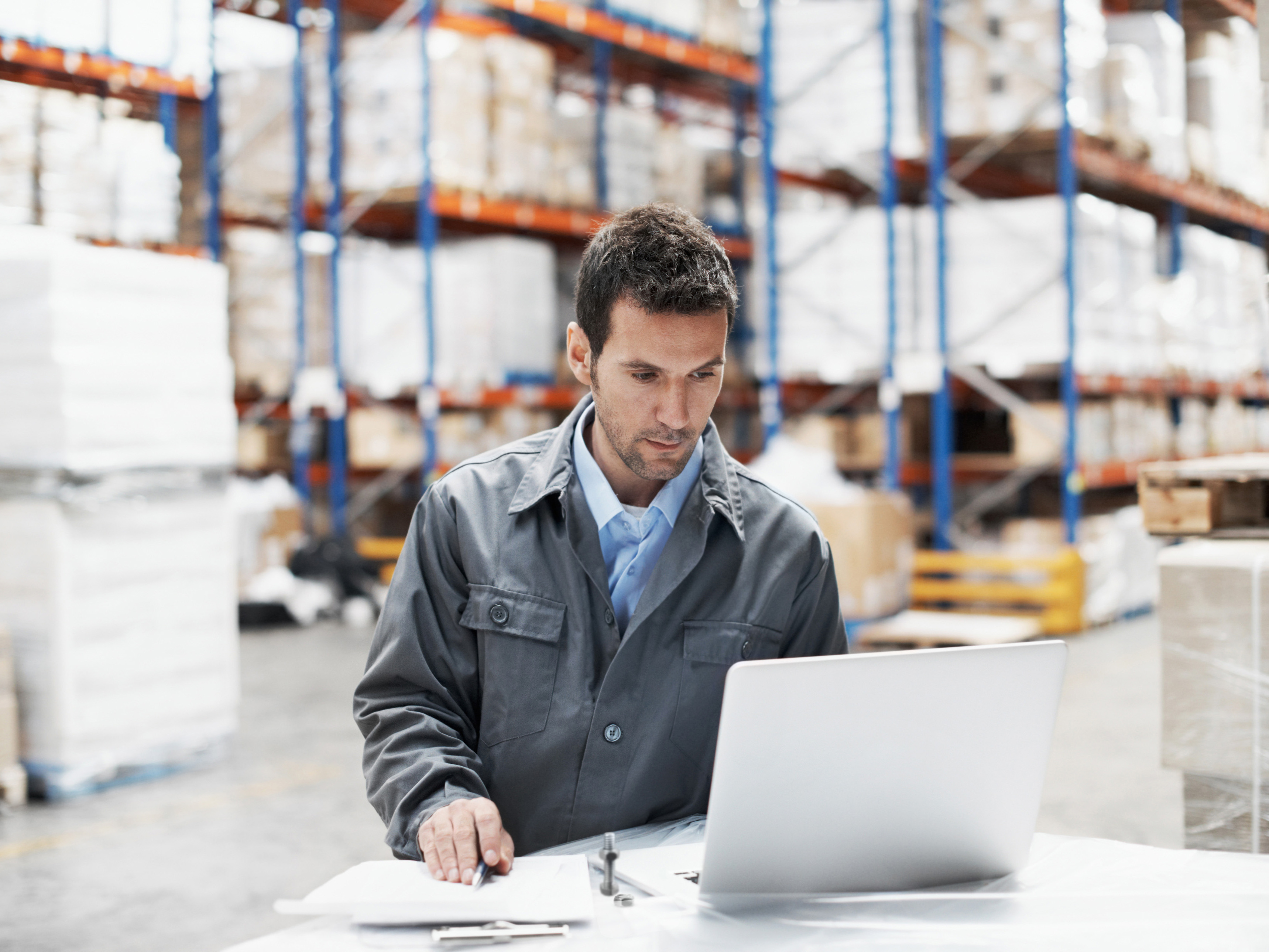 Man working at a laptop in a smart warehouse, with pallets and shelves in the background