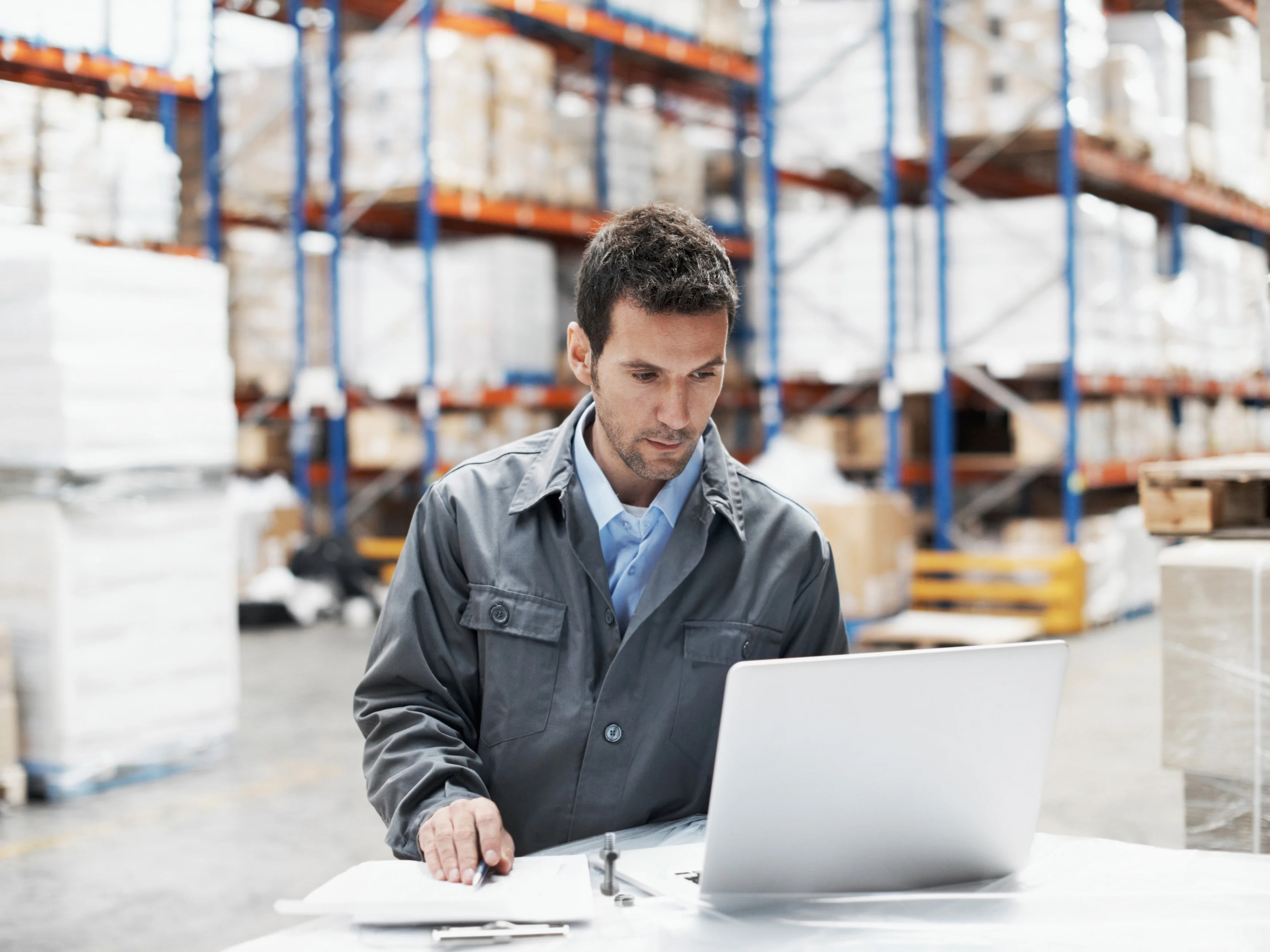 Man working at a laptop in a smart warehouse, with pallets and shelves in the background