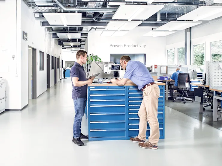 Two men in a test lab examine a sewing machine on a desk in an industrial lab with additional workstations in the background. Key visual for Expert Test Services.