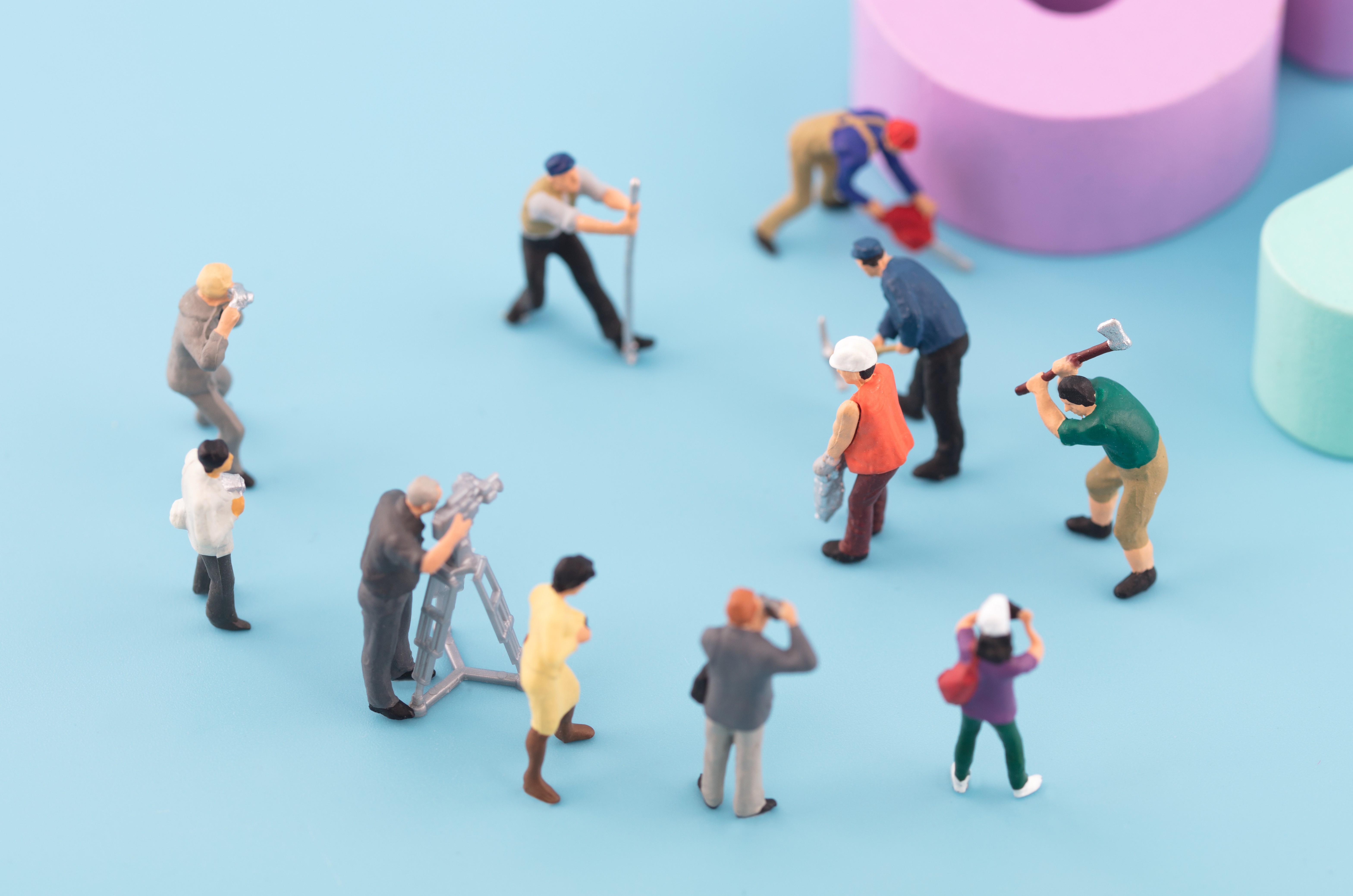 Miniature worker figures with tools and ladders on blue surface with large pastel purple and mint green objects.