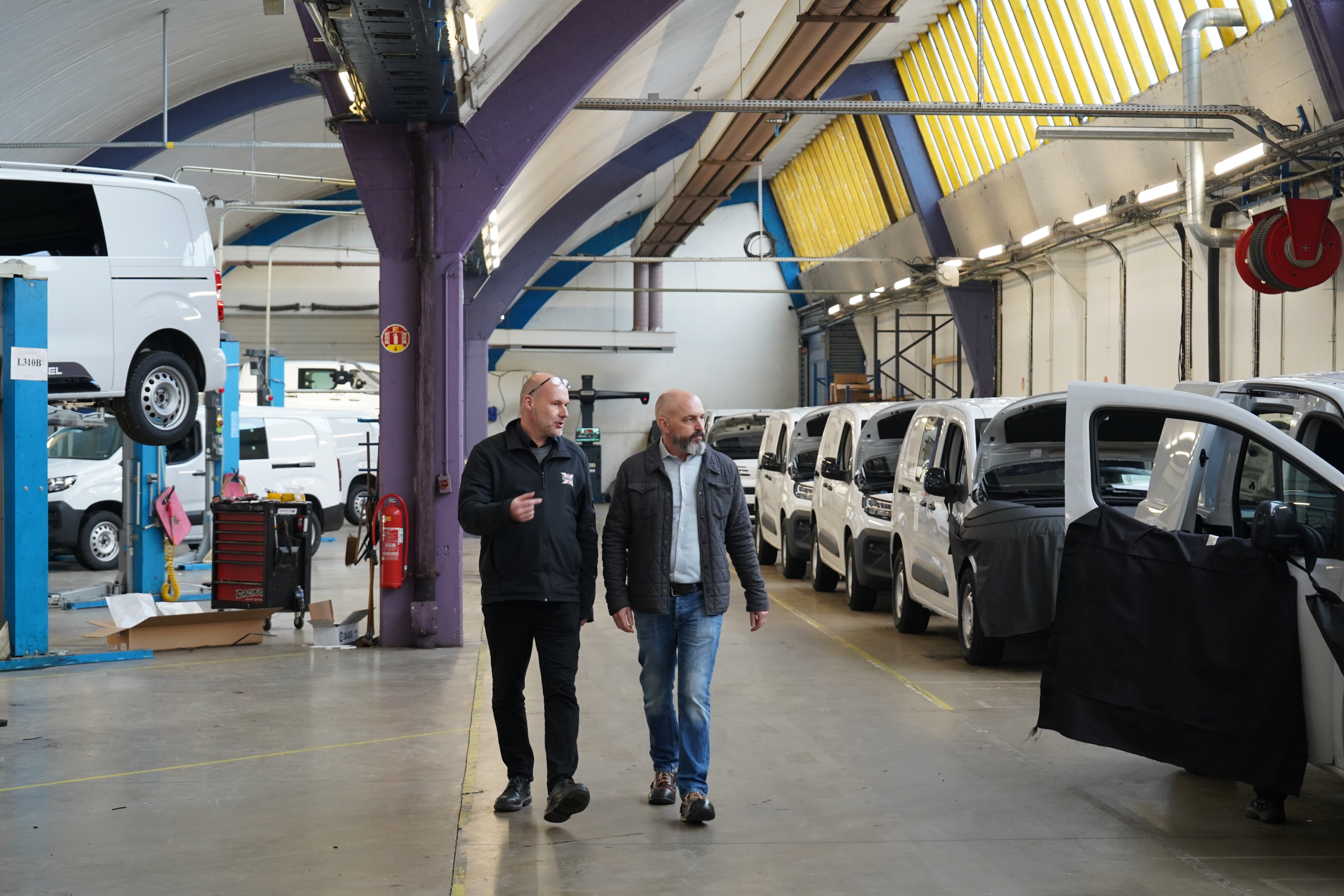 Two men walking in a factory hall with white cars in the background.