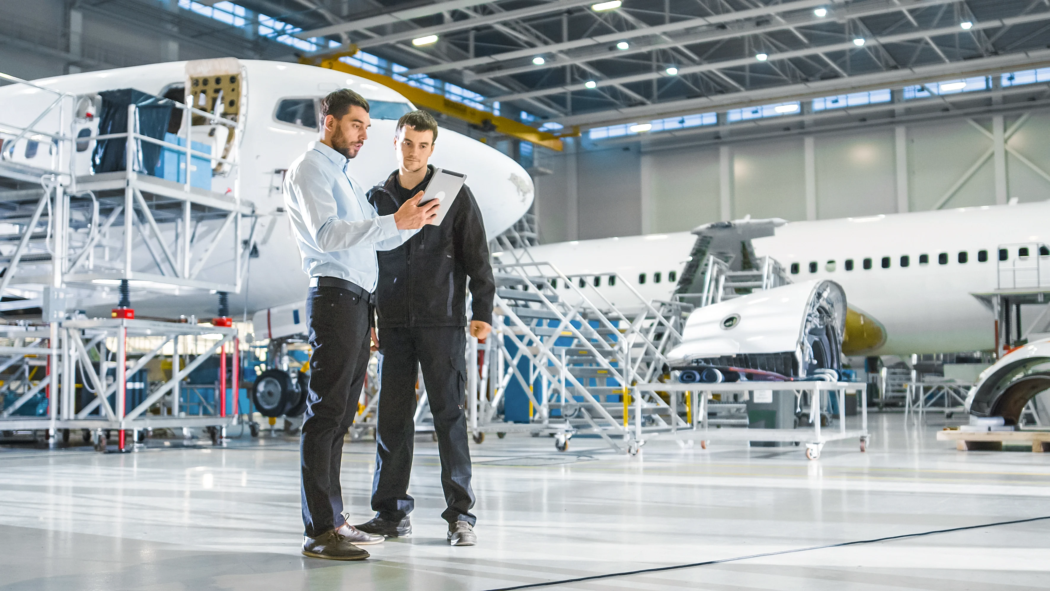 In an airplane factory, two men focus on a tablet, analyzing information while surrounded by aircraft components.