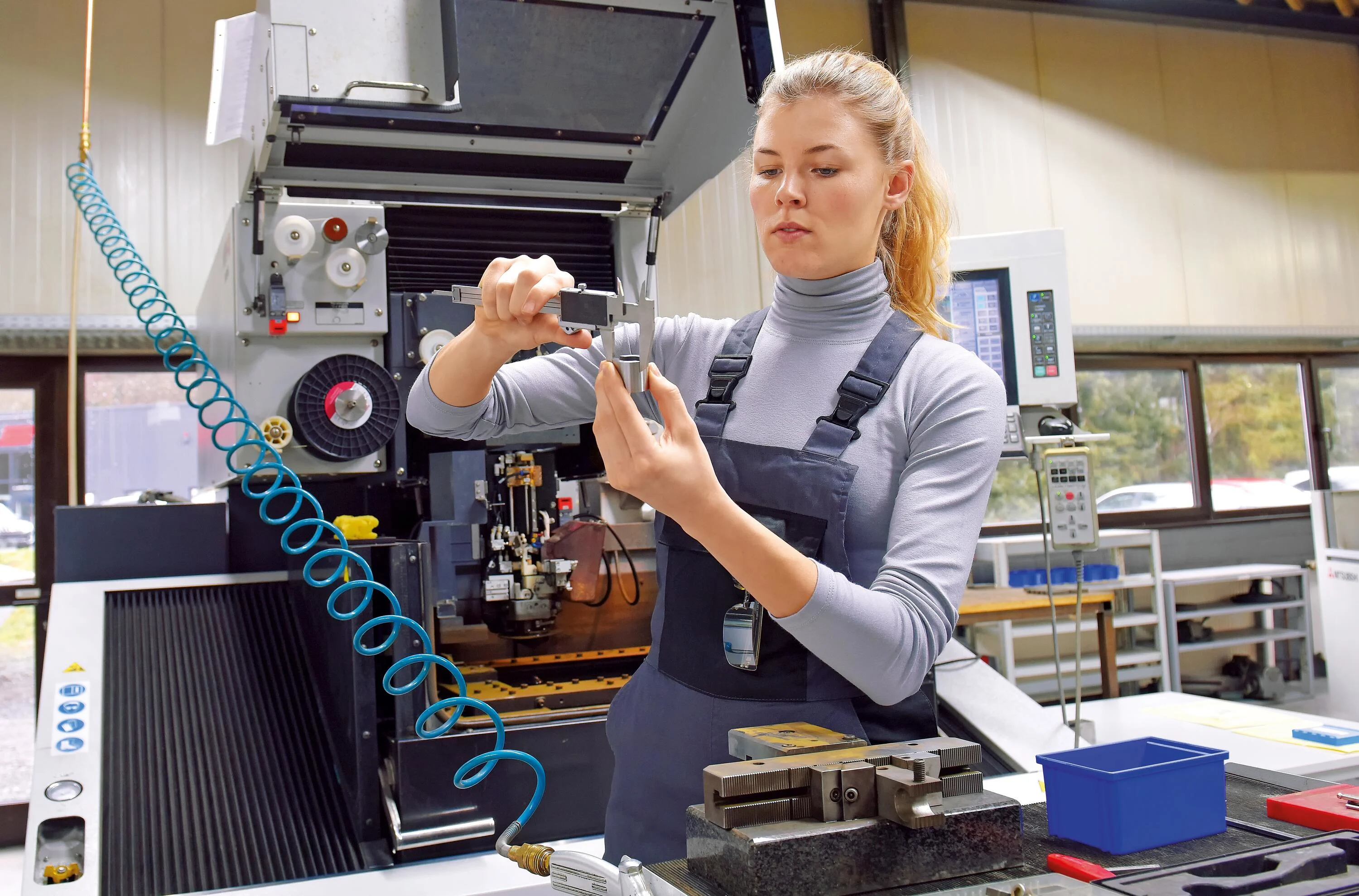 A young woman works as a computerized numerical control technician. She is seen at her workplace wearing a blue overall, measuring the outer diameter of a pipe.