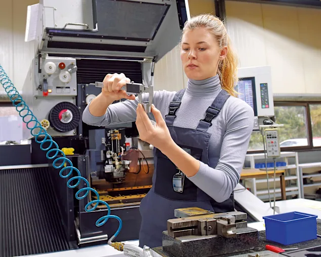 A young woman works as a computerized numerical control technician. She is seen at her workplace wearing a blue overall, measuring the outer diameter of a pipe.