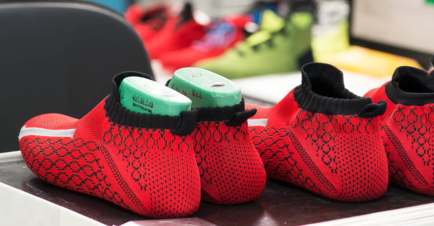 Red and black knitted shoe uppers with green molds inside, placed on a table. Background shows blurred colorful shoes.