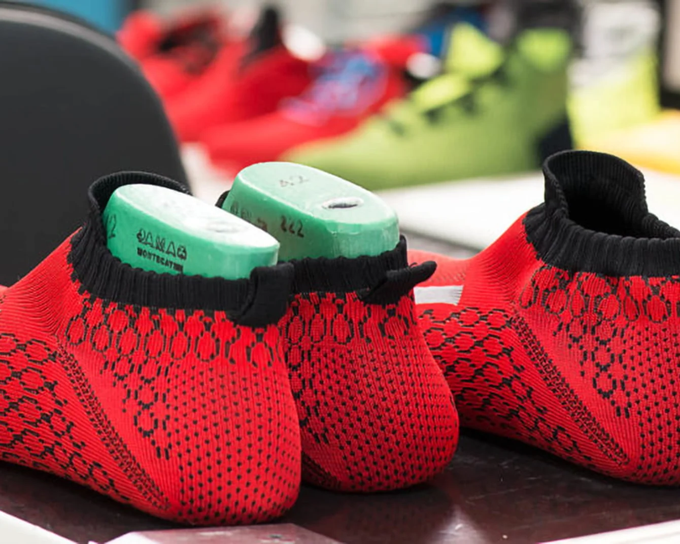 Red and black knitted shoe uppers with green molds inside, placed on a table. Background shows blurred colorful shoes.