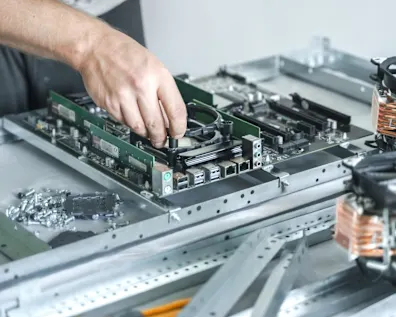 Technician's hand installing component on green computer motherboard surrounded by hardware parts and cooling fans on workbench