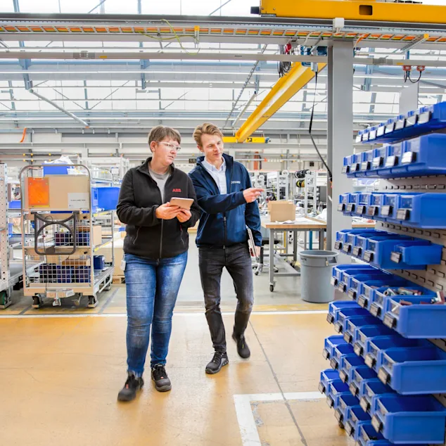 Two people walk through the shop floor, the woman holding a tablet in her hand, the man pointing to a mobile trolley equipped with SmartLabel Cloud bins.