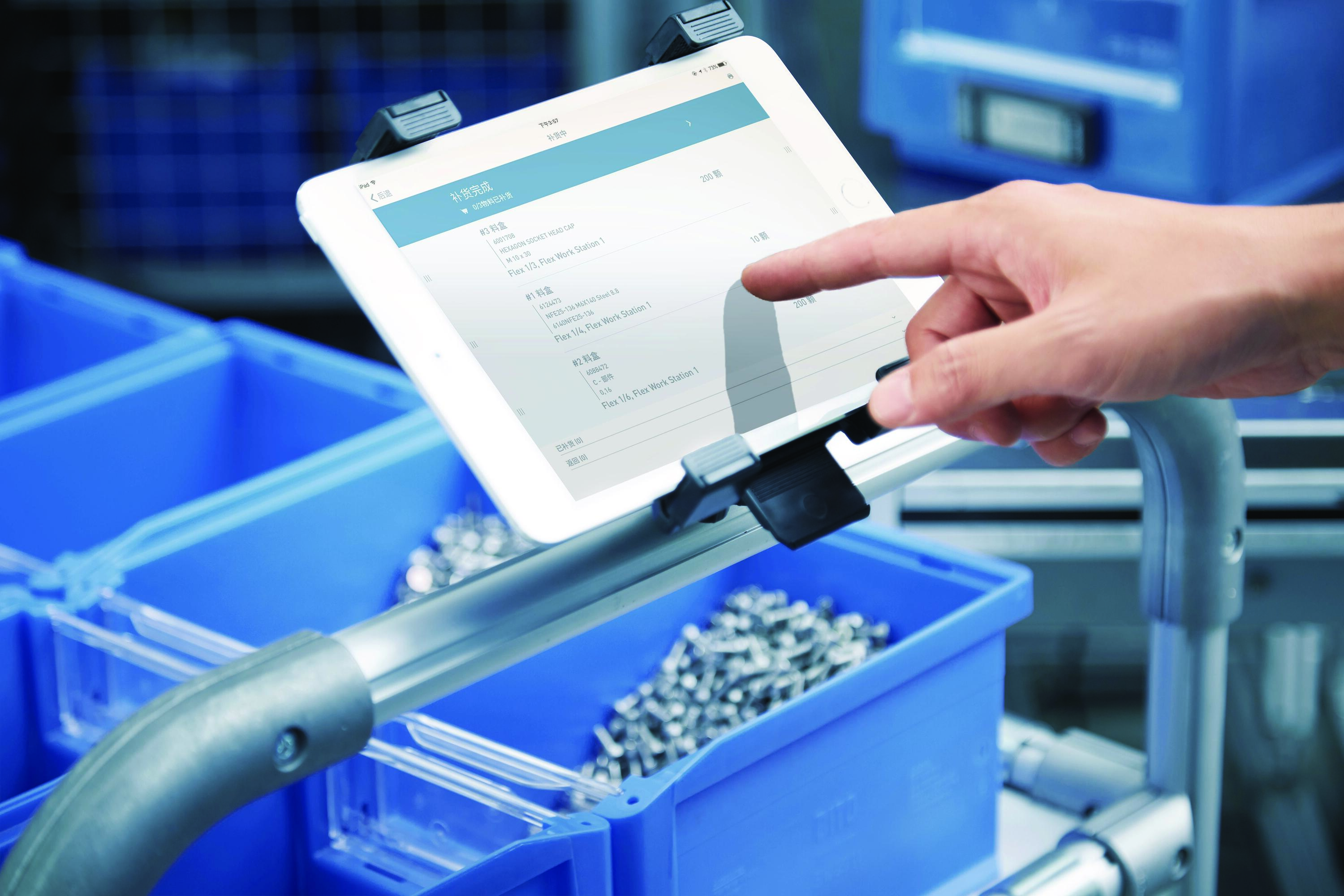 Hand pointing at white tablet mounted on stand displaying inventory data, with blue storage bins containing metal parts below.