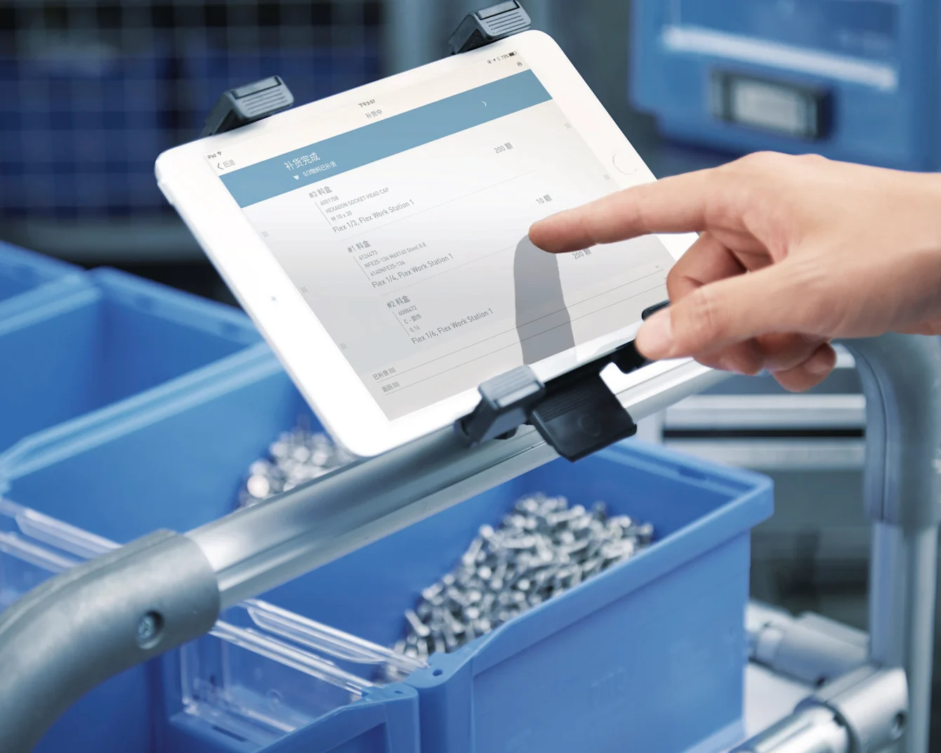 Hand pointing at white tablet mounted on stand displaying inventory data, with blue storage bins containing metal parts below.