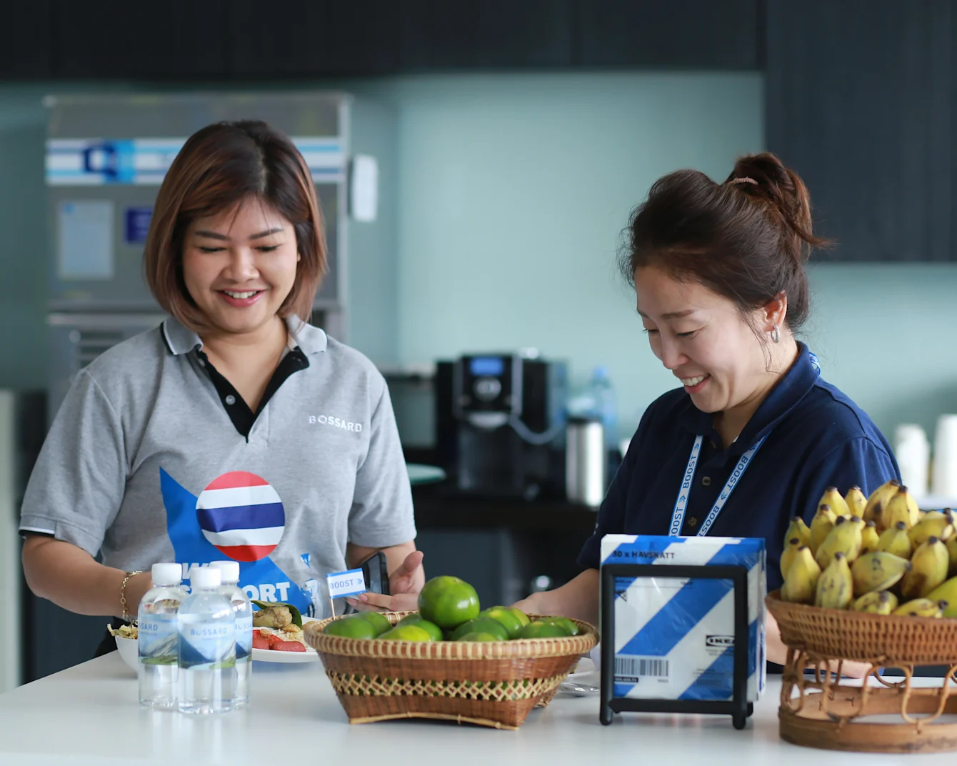 Two women smile joyfully while standing beside a table adorned with an assortment of fresh fruit.