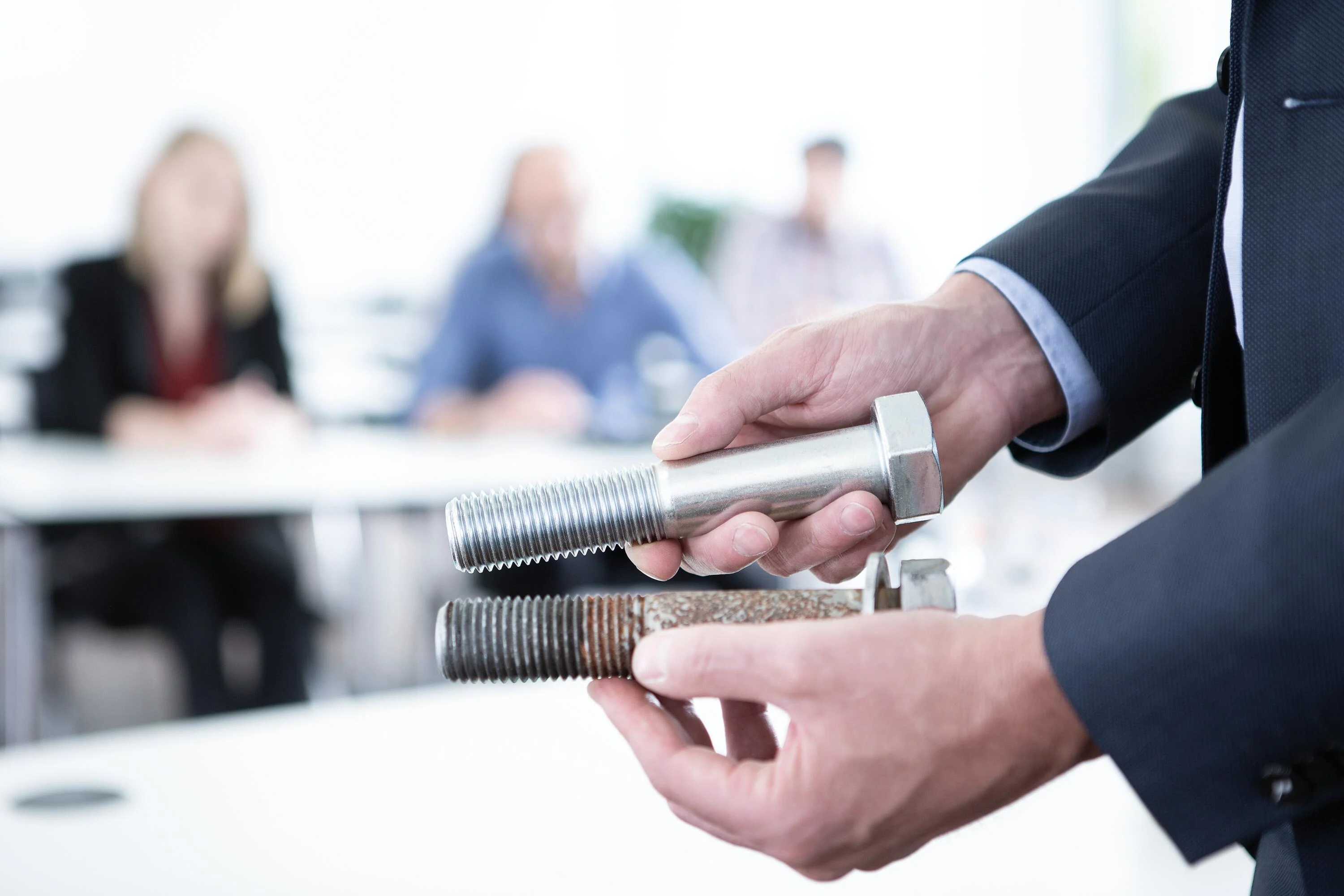Two hands hold bolts: the right a new, corrosion-free bolt; the left a corroded one. Three persons sit blurred in the background.