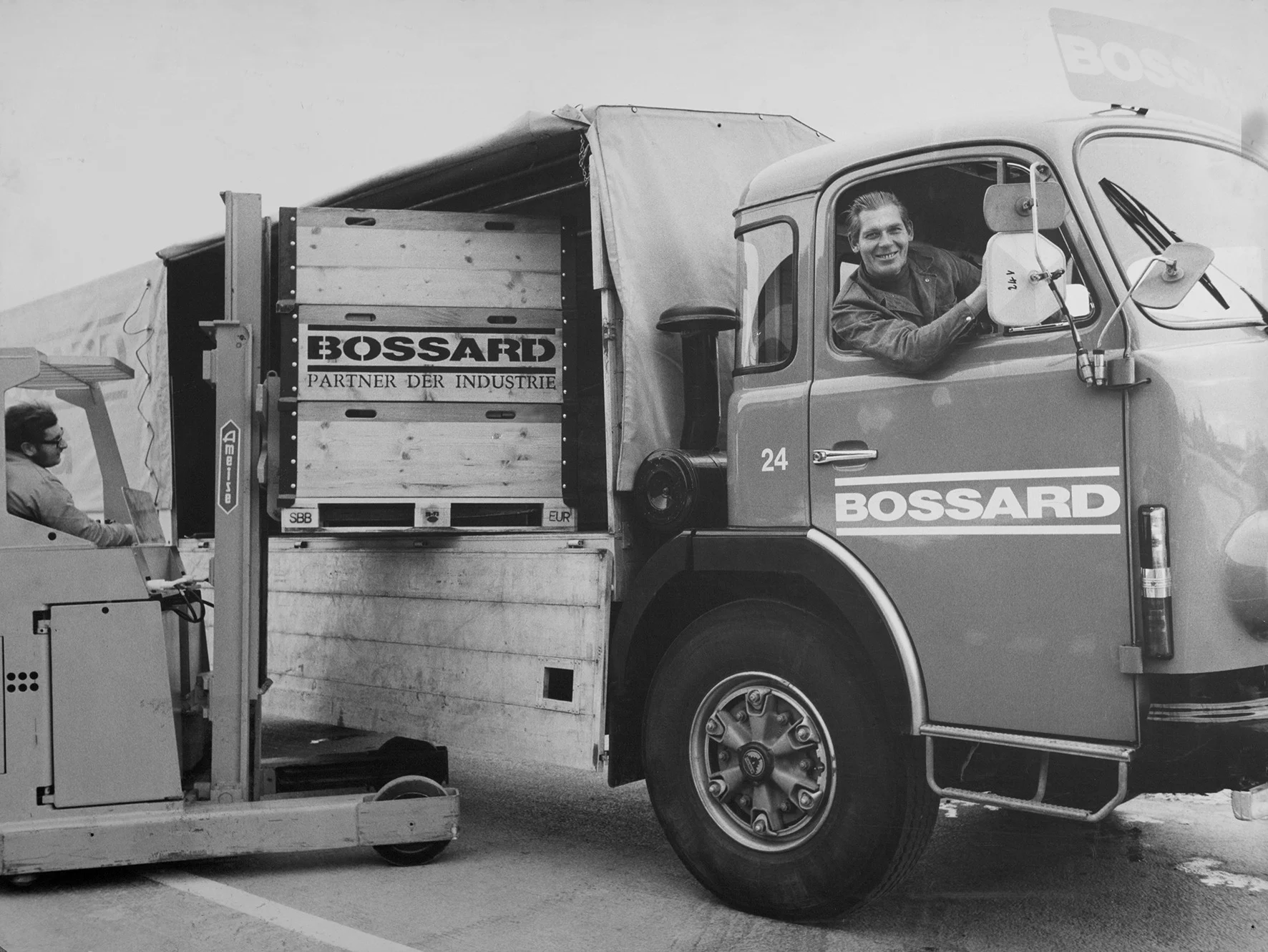 一张过去的黑白照片显示一名男子在堆垛机上卸载Bossard卡车,卡车内的另一名男子微笑,营造出合作的场景。