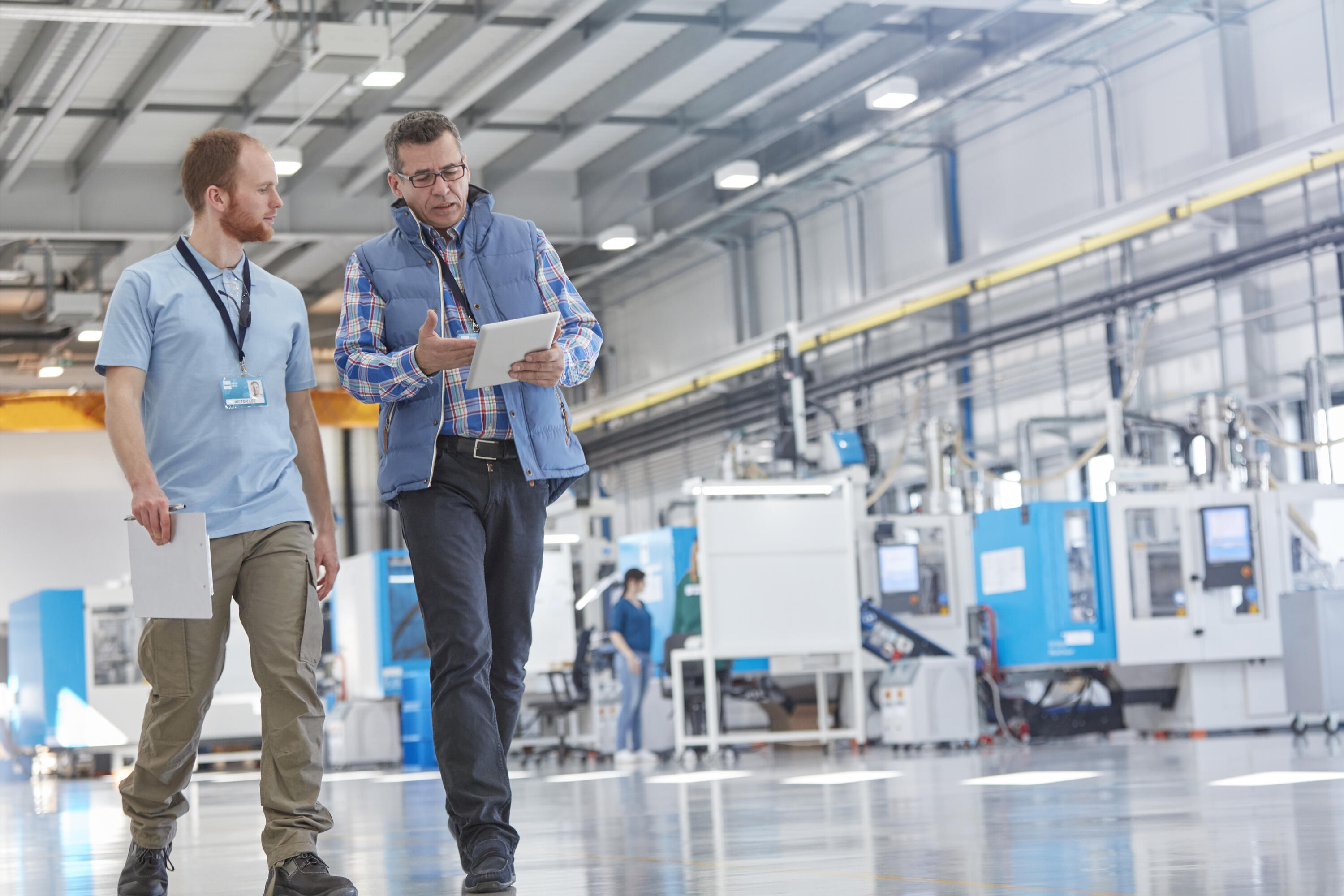 Two men walking through a customer site's production line. One holds a tablet, explaining to the other. Background shows the production/assembly line. Key visual for Expert Walk only.