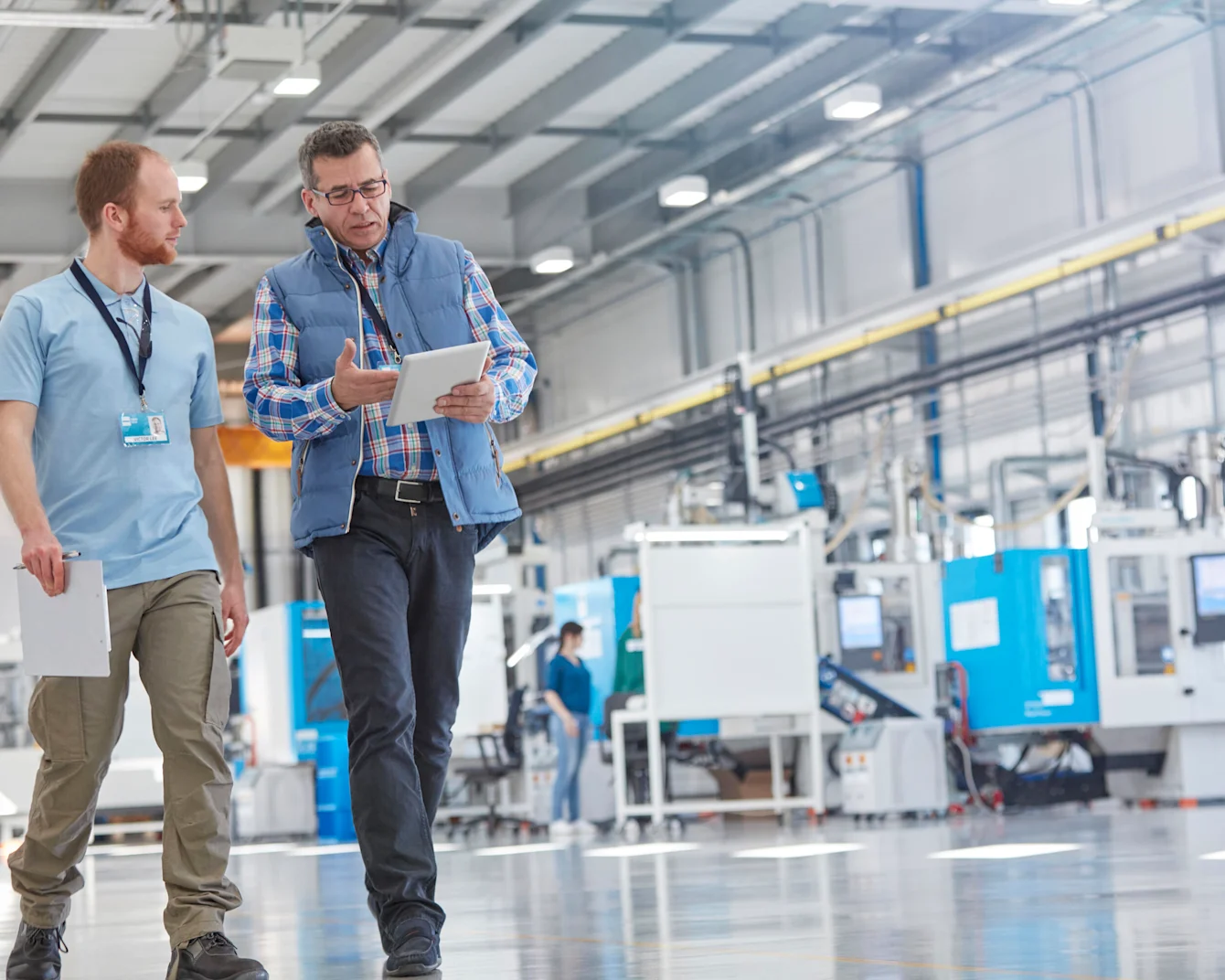 Two men walking through a customer site's production line. One holds a tablet, explaining to the other. Background shows the production/assembly line. Key visual for Expert Walk only.